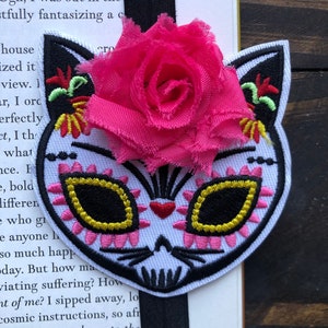 May include: A black and white embroidered cat bookmark with a pink flower on top. The cat has a sugar skull design with yellow and pink accents. The bookmark is attached to a black elastic band.