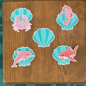 May include: Five embroidered patches featuring sea life and shells. One patch shows a pink crab on a teal shell, one a pink seahorse on a teal shell, one a pink dolphin on a teal shell, one a pink shark on a teal shell, and one a lone teal shell.