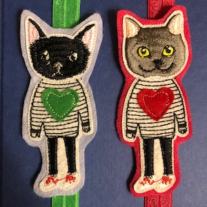 May include: Two felt cat bookmarks with striped shirts and hearts. The cat on the left has a green heart and the cat on the right has a red heart. Both cats are wearing black shoes.