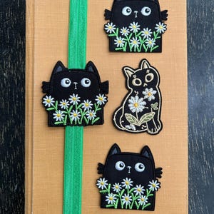 May include: Four black cat patches with white daisy accents are displayed on a tan book with a green elastic band. The cat patches have embroidered details and are in various poses. The background is a dark wooden surface.