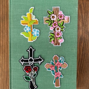 May include: Four embroidered cross patches with floral designs. One is gold with blue flowers, another is brown with pink flowers, a third is black with red roses, and the last is brown with red roses.
