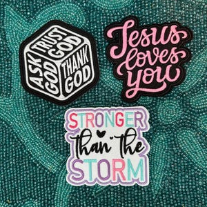 May include: Three embroidered patches with inspirational messages. One is a black and white hexagon with "Ask God, Trust God, Thank God". Another says "Jesus loves you" in pink script. The third says "Stronger Than the Storm".