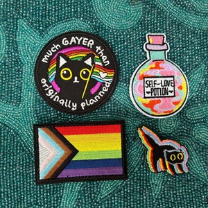 May include: A collection of embroidered patches featuring whimsical designs, including unicorns and a cat with a unicorn horn. The patches are in various colors, including pink, yellow, and blue, and are displayed on a blue background.