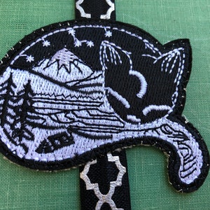 May include: Black and white embroidered cat bookmark with a mountain and stars design. The cat is curled up sleeping.