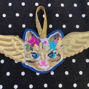 May include: A gold sequined cat with wings and a flower crown. The cat is on a blue background with a gold loop for hanging.