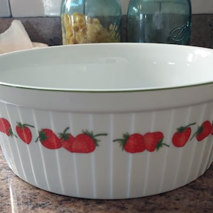 May include: A white ceramic mixing bowl with a ribbed exterior surface and a repeating pattern of red strawberries with green leaves encircling the middle. The rim of the bowl features a thin green line, and the interior is smooth and white.