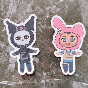 May include: Two cartoon character stickers. One is a black and white skull-faced character wearing a black and white outfit. The other is a pink bunny-eared character with a blue and white outfit.