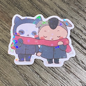 May include: Two cartoon characters wearing blue coats and red scarves. The character on the left has a skull face and cat ears. The character on the right has a round face and a gray hat. Both characters are standing side by side.