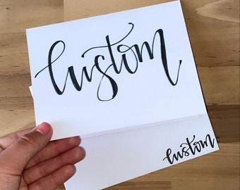 Puede incluir: Una tarjeta blanca con la palabra "custom" escrita en cursiva negra. La tarjeta está doblada por la mitad y la palabra "custom" es visible en ambos lados.