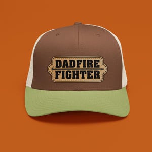May include: A brown and white trucker hat with a green brim. The hat features a rectangular patch with the words "DADFIRE FIGHTER" in a bold, vintage-style font. The hat is set against an orange background.
