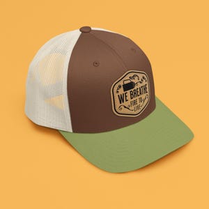 Könnte beinhalten: Eine braune und weiße Trucker-Kappe mit grünem Schirm. Die Kappe hat einen braunen Leder-Patch mit dem Text "WE BREATHE FIRE TO LIFE" in Schwarz.
