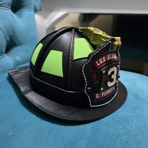 Pode incluir: Um capacete de bombeiro preto com detalhes retangulares em verde-limão brilhante na cúpula. O escudo frontal exibe o texto LOS ALAMOS, o número 3 e C. FLORES G. Um emblema de dragão dourado está fixado no topo do escudo frontal.