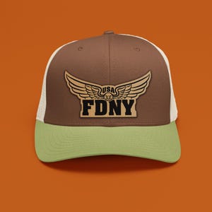 May include: A brown and green trucker hat with a white mesh back. The front features a leather patch with the text "FDNY" below a winged emblem and the word "USA".