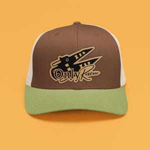 Puede incluir: Una gorra de camionero marrón y verde con una malla blanca en la parte posterior. La gorra presenta un gráfico en blanco y negro de un avión con el texto "Only Rescue" en marrón.