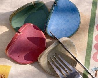 May include: Four ceramic ashtrays in shades of green, blue, pink, and beige, displayed on a patterned tablecloth. A silver fork and knife are positioned nearby, possibly for an outdoor dining arrangement.