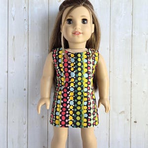 May include: A doll with long brown hair wearing a sleeveless dress featuring a black background with vertical stripes of multicolored polka dots in shades of pink, yellow, light blue, and green. The dress has a round neckline.