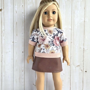 May include: A doll with blonde hair wearing a short-sleeved shirt featuring a white background with a floral pattern in shades of pink, brown, and black, accented by a light pink ribbed collar and hem, paired with a solid brown faux leather skirt.