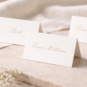 Könnte beinhalten: Elfenbeinfarbene Tischkarten mit goldener Kalligraphie, darunter "Bride" und Namen, auf einer Steinoberfläche mit weißen Blumen angeordnet. Die Karten sind gefaltet, um aufrecht zu stehen, ideal für Hochzeiten.