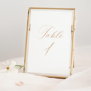May include: A gold-framed table number sign with the text "Table 1" in elegant script. A white flower and a gold ring are in the foreground. The sign is on a white background.
