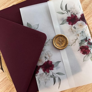May include: Wedding invitation suite featuring a burgundy envelope and a floral design with white and burgundy flowers. The invitation is sealed with a gold wax seal. The design includes watercolor-style flowers and greenery.