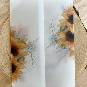 Op de afbeelding: Wit papier met twee panelen met aquarel zonnebloemen en groen. De zonnebloemen zijn geel en bruin, met groene bladeren. Het papier is in het midden gevouwen en de achtergrond is een lichtblauwe en witte wassing.