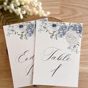 Op de afbeelding: Twee witte tafelkaarten met bloemen aquarel ontwerpen in blauw en wit. De kaarten hebben elegante kalligrafie met de tekst "End" en "Table 1". De kaarten liggen op een houten oppervlak met witte bloemen op de achtergrond.