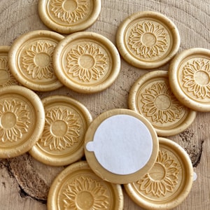 Könnte beinhalten: Goldfarbene Wachssiegel mit einem Sonnenblumenmuster. Die runden Siegel sind mit einem detaillierten Sonnenblumenmuster geprägt. Ein Siegel hat eine weiße Kleberückseite. Die Siegel sind auf einer Holzoberfläche angeordnet.