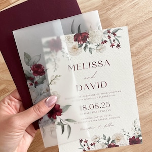 May include: Elegant wedding invitation suite. The design includes a burgundy envelope, a vellum overlay, and a textured invitation card. The invitation features the names "Melissa and David", the date "8.08.25", and floral details.