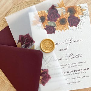 Puede incluir: Conjunto de invitación de boda con un sobre burdeos, una tarjeta adornada con girasoles y rosas burdeos, y una superposición translúcida. La invitación incluye los nombres "ssa Spencer and David Butler" y la fecha "16 de octubre de 2025". Un sello de cera dorado está presente.