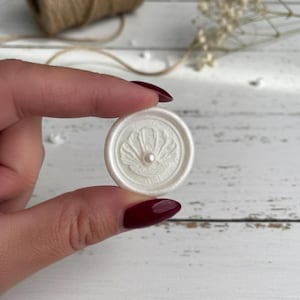 May include: A white wax seal featuring a pearl and seashell design. The round seal is held between fingers. A spool of twine and dried flowers are in the background.