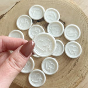 Könnte beinhalten: Eine Sammlung elfenbeinfarbener Wachssiegel, jedes mit einem Herz- und Flugzeugdesign geprägt. Die runden Siegel sind auf einer Holzoberfläche angeordnet. Ein Siegel wird von einer Hand mit lackiertem Nagel gehalten.