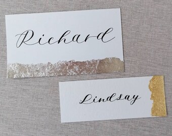 Könnte beinhalten: Zwei weiße Tischkarten mit eleganten kalligraphischen Namen. Eine Karte trägt den Namen "Richard" mit einem silbernen Folienakzent, die andere den Namen "Lindsay" mit einem goldenen Folienakzent. Die Karten liegen auf einer neutralen Leinenoberfläche.