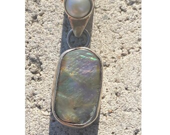 May include: Silver pendant featuring a pearlescent, rectangular stone with iridescent hues of green, blue, and purple. A small, round pearl is set at the top of the pendant. The pendant is displayed on a textured, gray surface.