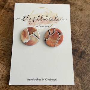 May include: A pair of round, brown and white patterned earrings with a black border. The earrings are displayed on a white card with the text "The Gilded Boho by Taryn Elsa" and "Handcrafted in Cincinnati".