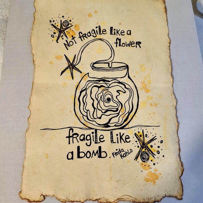 Puede incluir: Obra de arte dibujada a mano sobre papel envejecido. Un dibujo lineal negro representa una bomba con una rosa en su interior, conectada al texto "Not fragile like a flower" y "fragile like a bomb". La firma del artista, "Frida Kahlo", est&aacute; presente.