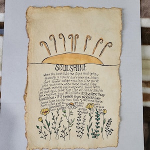Pu&ograve; includere: Un'illustrazione disegnata a mano su carta pergamena con un sole giallo e fiori. Il testo dice "SOULSHINE" e include una citazione sul trovare la luce nei momenti bui.