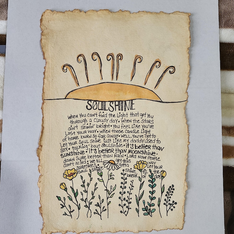 Pu&ograve; includere: Un'illustrazione disegnata a mano su carta pergamena con un sole giallo e fiori. Il testo dice "SOULSHINE" e include una citazione sul trovare la luce nei momenti bui.