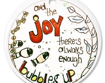 Op de afbeelding: Een witte, ronde keramische ornament met de tekst "and the joy there's always enough bubbles up" in een handgetekende stijl. Het woord "joy" is rood, en het ornament bevat illustraties van vissen en planten.