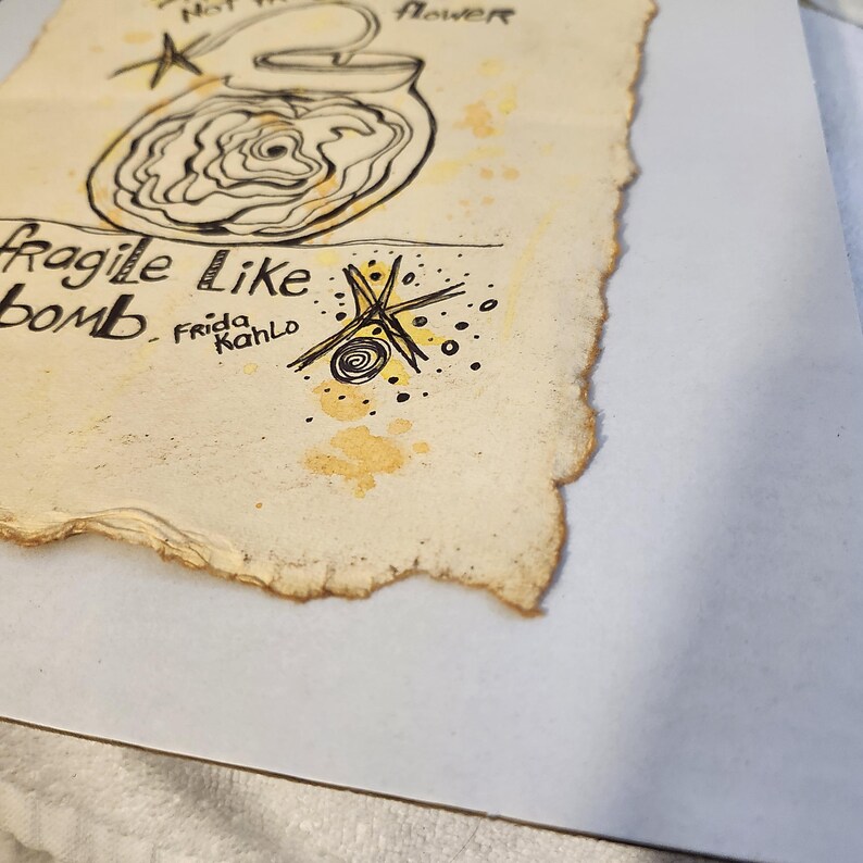 Puede incluir: Obra de arte dibujada a mano sobre papel envejecido que presenta un tarro con un dise&ntilde;o floral, acompa&ntilde;ado del texto "Not the flower" y "Fragile Like bomb Frida Kahlo." La obra utiliza tinta negra y detalles amarillos.