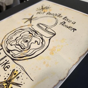 Puede incluir: Obra de arte dibujada a mano sobre papel envejecido, con un dise&ntilde;o en forma de bomba con un interior floral. El dibujo a tinta negra incluye el texto "Not fragile like a flower". El papel tiene manchas amarillas-marrones y est&aacute; enmarcado.