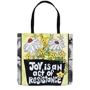 May include: A yellow and white tote bag with a black handle. The bag features a hand-drawn illustration of flowers in a pot with the text "JOY is an act of resistance".