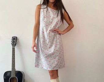 May include: A sleeveless, light blue dress with a small red floral pattern. The dress is paired with cream-colored knee-high boots. A black acoustic guitar leans against a white wall.