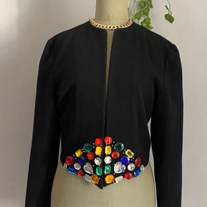 Puede incluir: Chaqueta negra con un dise&ntilde;o con incrustaciones de joyas. La chaqueta presenta un dise&ntilde;o sin cuello y mangas largas. Los adornos de joyas son de varios colores, incluyendo rojo, azul, verde y amarillo. Un collar de cadena dorada rodea el cuello del maniqu&iacute;.