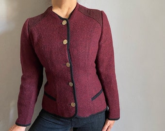 May include: A burgundy wool jacket with a round collar, black trim, and gold buttons. The jacket has a tailored fit with a quilted shoulder detail and two front pockets. The sleeves are long, and the jacket is buttoned up.