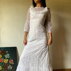 Op de afbeelding: Een witte maxi-jurk met een transparante overlay en kantversiering. De jurk heeft een ronde hals, 3/4 mouwen en een gelaagde rok.