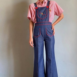 Può includere: Salopette in denim blu con finiture rosse e ricami floreali. La salopette ha vita alta e gambe ampie. L'outfit include una camicia a maniche corte a quadri rossi e bianchi. Le bretelle hanno bottoni rossi.