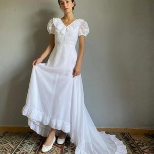 Puede incluir: Un vestido de novia blanco vintage con un dobladillo alto-bajo y una larga cola. El vestido presenta un escote con ribete de encaje, mangas cortas abullonadas y un corpiño ajustado. El vestido está hecho de una tela ligera, adecuado para una boda.