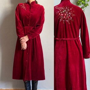 May include: A long, button-down, red velvet robe with long sleeves and a matching belt. The robe features floral embroidery on the upper chest and back. The robe is knee-length and has a high collar.
