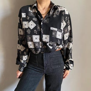 May include: Black button-down shirt with a patchwork pattern of white squares, floral designs, and geometric shapes. The shirt has long sleeves and a collared neckline. The model is wearing dark wash denim jeans.