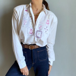 Könnte beinhalten: Weißes Button-Down-Hemd mit gestickten Aufnähern mit einem Haus, Teddybären und dem Text "FOR GET ME NOT". Das Hemd hat lange Ärmel und wird mit einer dunkelblauen Jeans und einem braunen Gürtel kombiniert.